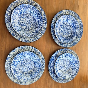 May include: Four sets of round dinnerware, each consisting of a larger plate and a smaller bowl. The pieces feature a blue and white speckled enamel pattern with a dark rim. The set is arranged in a two-by-two grid on a flat surface.