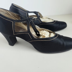 May include: A pair of black satin T-strap heels featuring a rounded toe, a mid-height curved heel, and delicate tan stitching along the edges. The shoes have a cutout design on the sides and a central strap that connects to the collar.