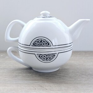 May include: White ceramic teapot and teacup set with black Celtic knot designs. The teapot has a lid and spout, and the teacup is integrated into the base. The set is on a wooden surface.