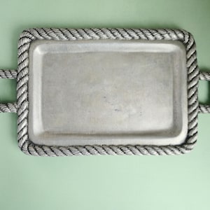 May include: A rectangular silver-toned metal tray featuring a textured rope-style border and two matching rope-detailed handles on the sides. The central surface of the tray is smooth and flat, framed by the raised, twisted rope pattern.