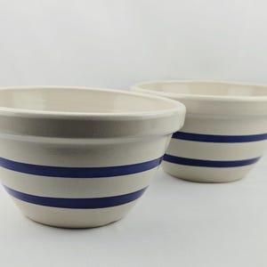 May include: Two cream-coloured ceramic mixing bowls featuring two horizontal navy blue stripes around the exterior. Each bowl has a wide, rounded rim and a smooth, glossy finish, presented against a plain, light-coloured background.