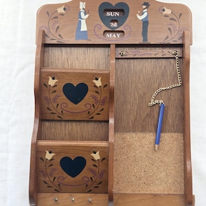 Puede incluir: Organizador de pared de madera con calendario, dos ranuras para correo, un corcho y ganchos para llaves. El organizador presenta diseños florales y de corazón pintados, con un lápiz azul colgando. El calendario muestra domingo 24 de mayo.