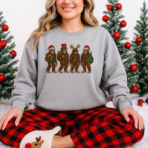 Peut inclure: Un sweat-shirt gris avec un motif festif de quatre personnages de Bigfoot vêtus de tenues de fête. Les personnages de Bigfoot sont bruns et portent des chapeaux de Père Noël, un haut-de-forme et des bois. La personne porte un pantalon de pyjama à carreaux rouges et noirs.