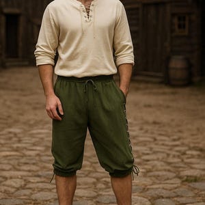 May include: A person wearing a cream-colored long-sleeve shirt with a laced neckline and olive green knee-length shorts. The shorts have a drawstring waist and decorative lacing on the sides. The background features wooden buildings.