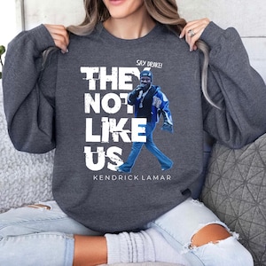 May include: A dark gray sweatshirt with a white graphic print that says "They Not Like Us" with a picture of a man in a blue and white jacket. The text "Say Drake!" is above the graphic and "Kendrick Lamar" is below the graphic.