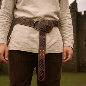 May include: A brown leather belt with Celtic knotwork design. The belt features a brass buckle and is worn over a cream-coloured long-sleeved shirt and brown trousers. The belt is approximately 3.8 cm wide.