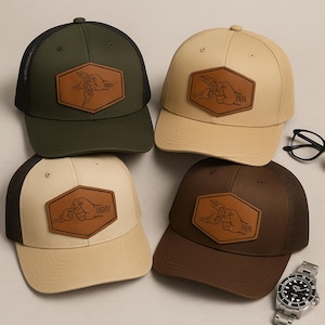 Może przedstawiać: Cztery czapki bejsbolowe z siateczkowymi plecami i skórzanymi naszywkami. Czapki są w kolorze oliwkowym, beżowym i brązowym. Skórzane naszywki przedstawiają wzór pięści z napisami "DADDY" lub "PAPA".