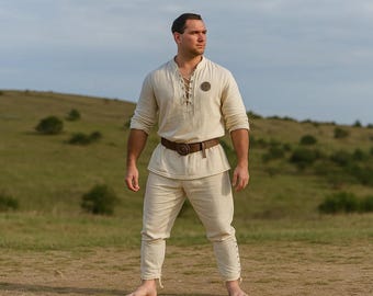 May include: A cream-colored linen outfit consisting of a long-sleeved shirt with a laced neckline and a pair of matching pants. The outfit is accessorized with a brown leather belt. The person is standing outdoors on a dirt path.