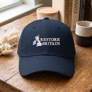 Może przedstawiać: Granatowa czapka z daszkiem z białym napisem "RESTORE BRITAIN". Czapka posiada biały haft zarys Wysp Brytyjskich. Czapka leży na drewnianej powierzchni.