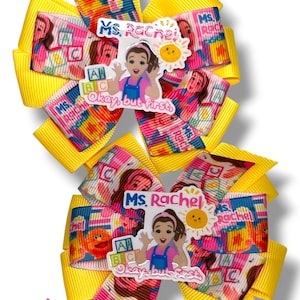 May include: Two hair bows featuring yellow ribbon loops and a central printed ribbon pattern showing Ms. Rachel, alphabet blocks, and a sun. Each bow has a central acrylic charm depicting Ms. Rachel with the text Ms. Rachel and Okay, but first.
