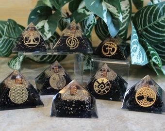 May include: A collection of black orgone pyramids with gold-colored symbols, including the Om symbol, a tree of life, and a yin-yang design. The pyramids are displayed on a clear stand with green foliage in the background.