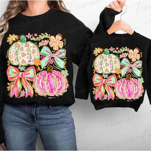 May include: Black sweatshirts with colorful pumpkin designs. The adult sweatshirt features a large pumpkin, bow, and floral design. The child's sweatshirt has a similar design. The pumpkins are white, pink, and orange with green and pink bows.