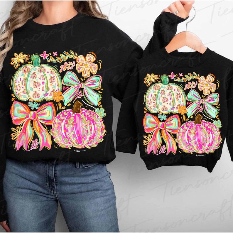 May include: Black sweatshirts with colorful pumpkin designs. The adult sweatshirt features a large pumpkin, bow, and floral design. The child's sweatshirt has a similar design. The pumpkins are white, pink, and orange with green and pink bows.
