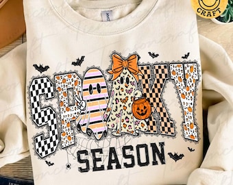 May include: A cream-colored crewneck sweatshirt featuring the text SPOOKY SEASON. The word SPOOKY is decorated with black and white checkerboard, leopard print, and striped patterns, while two ghosts and a pumpkin bucket are displayed.