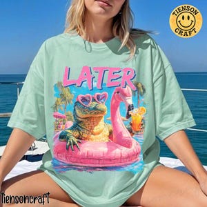 May include: Mint green t-shirt with a graphic featuring a cartoon alligator wearing heart-shaped sunglasses and a pink flamingo in a pool. The word "LATER" is printed above the graphic in pink. The shirt has a crew neck and short sleeves.