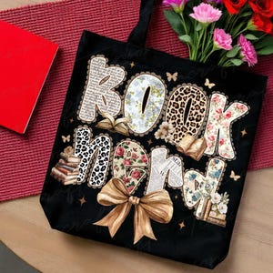 Puede incluir: Bolso tote negro con las palabras "BOOK NERD" en una variedad de fuentes decorativas, incluyendo floral, estampado de leopardo y peri&oacute;dico. Un gran lazo dorado en la parte inferior. Mariposas y peque&ntilde;as estrellas est&aacute;n dispersas alrededor del texto.
