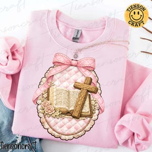 May include: A light pink sweatshirt featuring a detailed graphic of a cross, an open book, and a pink bow. The design is set within a quilted oval frame with floral accents. The brand name "Tiensoncraft" is visible.