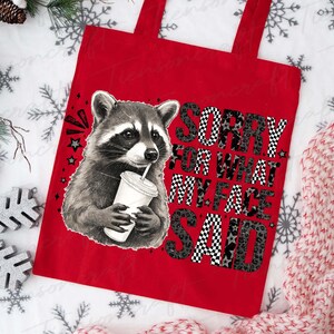 May include: Red tote bag featuring a raccoon drinking from a cup. The bag has the text "SORRY FOR WHAT MY FACE SAID" in a mix of checkered and leopard print patterns. Stars and other graphics are also included.