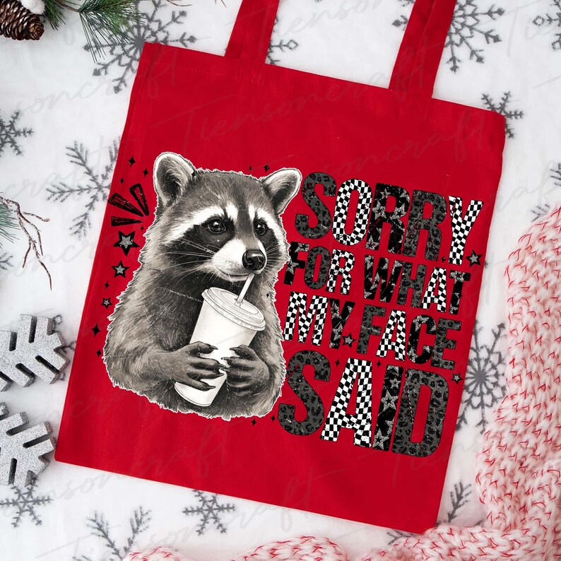 May include: Red tote bag featuring a raccoon drinking from a cup. The bag has the text "SORRY FOR WHAT MY FACE SAID" in a mix of checkered and leopard print patterns. Stars and other graphics are also included.