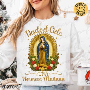 May include: White sweatshirt with gold embroidered text and a religious image. The image features a woman in a blue robe with gold accents, surrounded by a gold halo and roses. The text reads "Desde el Cielo Una Hermosa Mañana."