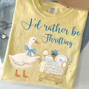 Op de afbeelding: Lichtgele t-shirt met de blauwe tekst "I'd rather be Thrifting". Een cartoon gans met een blauwe strik duwt een winkelwagentje vol vintage items, waaronder borden en een lamp.