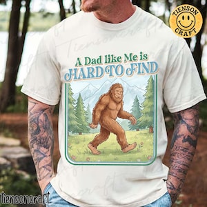 Pode incluir: Camiseta creme com uma ilustração de Bigfoot caminhando por uma cena de floresta. O texto "A Dad like Me is HARD TO FIND" é impresso acima da imagem em azul e verde. A camisa tem mangas curtas.