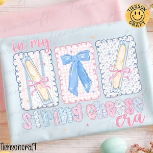 Könnte beinhalten: Hellblaues T-Shirt mit dem Text "in my string cheese era". Das Design zeigt drei gerahmte Illustrationen: zwei Käsesnacks mit rosa Schleifen und eine blaue Schleife. Das Design hat eine skurrile, pastellfarbene Ästhetik.