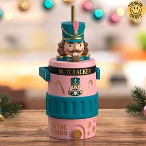 May include: A pink and teal cylindrical beverage dispenser featuring a nutcracker figure lid with a gold straw. The body displays the text NUTCRACKER and TIS THE SEASON, with candy cane and snowflake graphics. A small spigot is at the base.