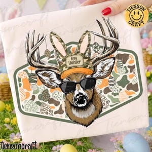 May include: A white t-shirt featuring a deer wearing sunglasses, a camouflage hat with the text "Egg Hunter", and bunny ears. The deer's antlers have hanging Easter eggs. The background has a green and orange camouflage pattern with more Easter eggs.