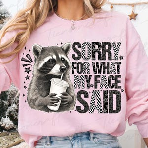 May include: Pink sweatshirt featuring a raccoon holding a drink with a straw. The text "SORRY FOR WHAT MY FACE SAID" is printed in a mix of checkered and leopard print patterns. Black stars and accents surround the raccoon and text.