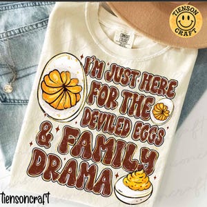 Può includere: T-shirt color crema con la scritta "I'M JUST HERE FOR THE DEVILED EGGS & FAMILY DRAMA" in marrone e bianco. La maglietta presenta illustrazioni di uova ripiene. È visibile un'etichetta Comfort Colors.