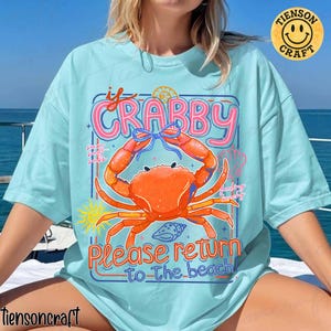 May include: Light blue t-shirt featuring a large orange crab illustration with the text "If Crabby Please Return to the Beach". The design includes a blue bow, seashells, and a sun. The shirt has a relaxed fit.