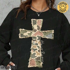 May include: Black sweatshirt featuring a camouflage cross design with frayed edges. The cross incorporates shades of green, brown, and pink. The words "He is Risen" are written in white script. A yellow smiley face logo is in the upper right corner.