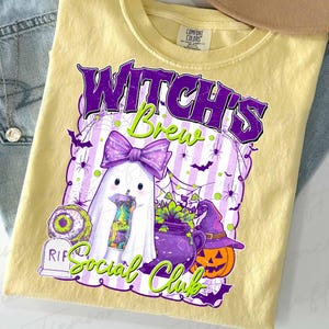 May include: A light yellow t-shirt featuring a Halloween-themed graphic. The design includes a ghost with a purple bow, a cauldron, a jack-o'-lantern, and the text "Witch's Brew Social Club" in purple and green.