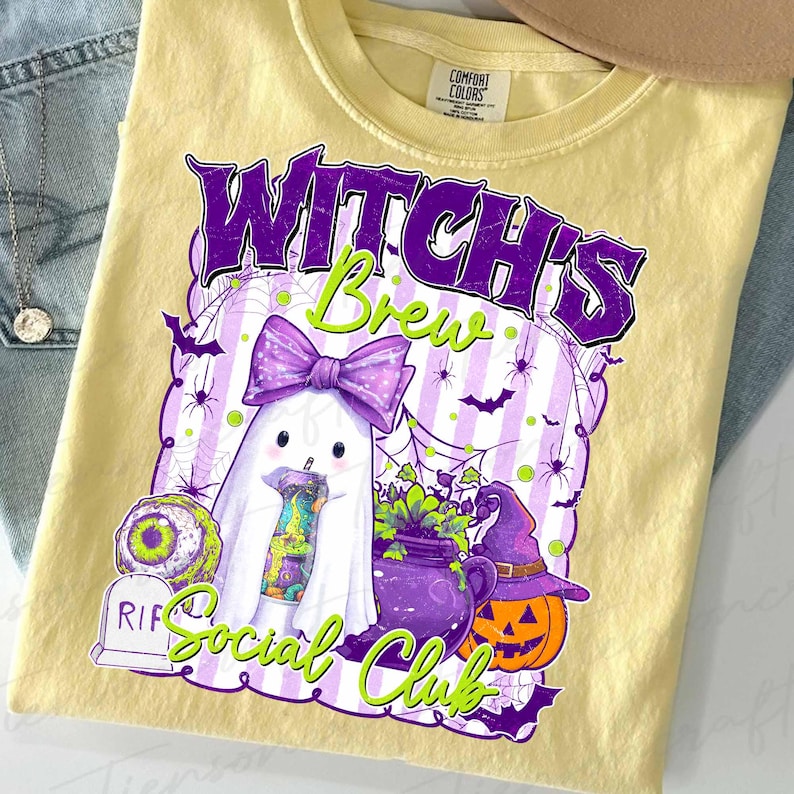 May include: A light yellow t-shirt featuring a Halloween-themed graphic. The design includes a ghost with a purple bow, a cauldron, a jack-o'-lantern, and the text "Witch's Brew Social Club" in purple and green.