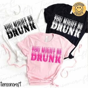 Puede incluir: Tres camisetas, blanca, negra y rosa, cada una con el texto "YOU MIGHT BE DRUNK" en una fuente audaz y en capas. Las camisetas blancas y negras tienen texto en blanco y negro, mientras que la camiseta rosa tiene texto rosa.