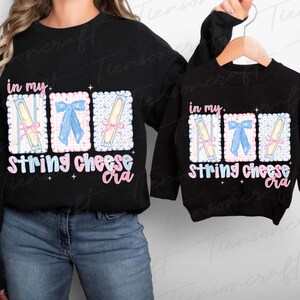 May include: Black sweatshirts with the text "in my...String cheese cra" in a whimsical font. The design features three illustrated rectangles with string cheese and bows. One sweatshirt is adult size, the other is child size.