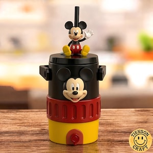 May include: A cylindrical beverage cooler featuring a black top section with a molded Mickey Mouse face, a red middle band, and a yellow base with a small spigot. A Mickey Mouse figurine sits on the lid, which includes a black straw attachment.