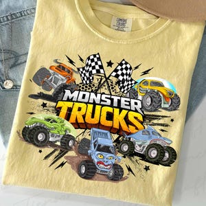 May include: Pale yellow t-shirt with a vibrant graphic of monster trucks. The design showcases several monster truck illustrations, chequered flags, and the text "MONSTER TRUCKS" in bold, orange and yellow letters.