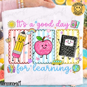 May include: A white t-shirt featuring a colorful graphic with the text It's a good day for learning. The design includes cartoon illustrations of a pencil, an apple, and a composition notebook, each inside a scalloped frame, alongside a school bus, globe, and small stars.