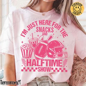 May include: Pale pink t-shirt featuring a darker pink graphic of a football, helmet, popcorn, and a drink. The text reads "I'M JUST HERE FOR THE SNACKS AND HALFTIME SHOW."