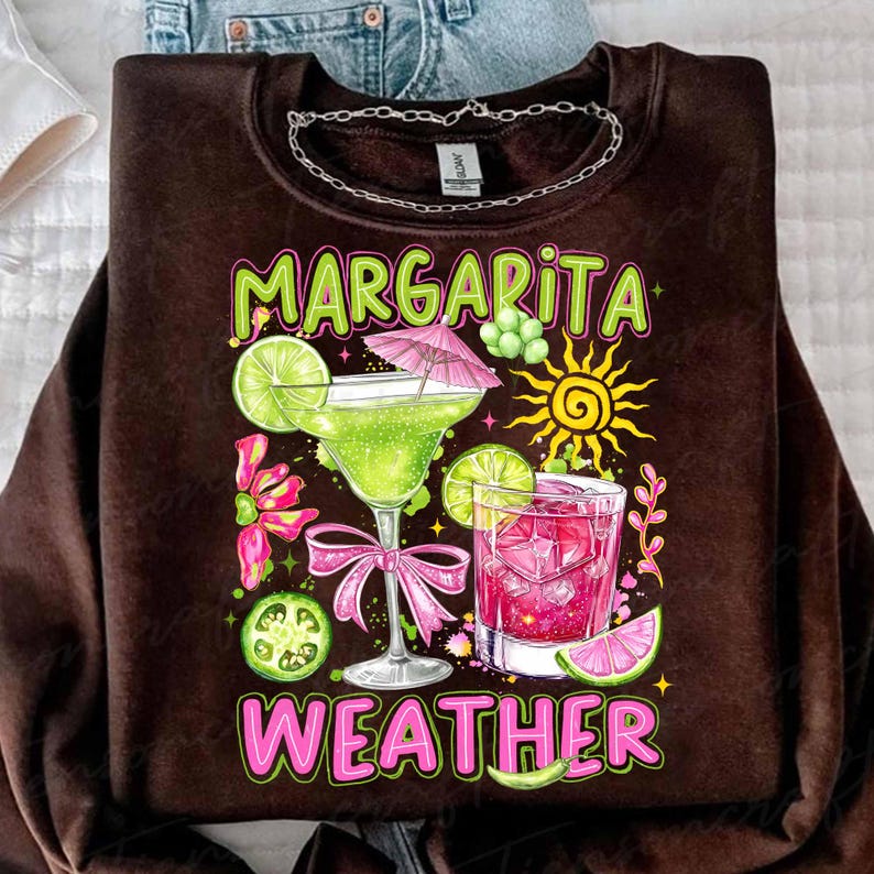 May include: Brown sweatshirt with a colorful graphic that reads "Margarita Weather." The design features two cocktails, lime slices, a sun, and pink accents. A silver chain is at the neckline.