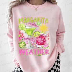 May include: Pink sweatshirt with a colorful graphic of two cocktails, lime slices, and the words "Margarita Weather" in green and pink. The design includes a margarita glass with a pink bow and a drink with ice cubes.