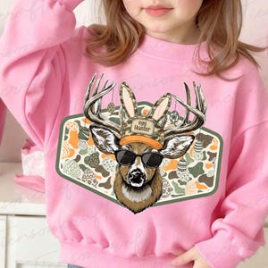 May include: Pink sweatshirt featuring a deer graphic wearing sunglasses, a camouflage hat with "Egg Hunter" text, and bunny ears. The graphic is set against a background of Easter eggs and a green border.