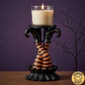 May include: A black candle holder featuring two crossed legs wearing black boots and orange and black striped stockings. The base is ruffled, and the top holds a clear glass jar containing a cream-colored candle with a lit wick.