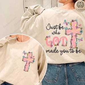 May include: Cream-colored sweatshirt featuring a pink cross adorned with flowers and butterflies. The text "Just be who God made you to be" is printed on the back. The design is a Christian-themed graphic.