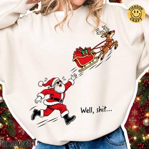 Könnte beinhalten: Ein cremefarbenes Sweatshirt mit einer Grafik, die den Weihnachtsmann zeigt, wie er einem roten, mit Geschenken beladenen Schlitten hinterherläuft, der von einem Rentier mit Sonnenbrille gezogen wird. Der Text "Well, shit..." steht in Schwarz.