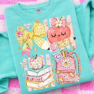 Pu&ograve; includere: Una felpa girocollo verde menta con una stampa grafica raffigurante un grande fiocco a quadretti gialli, una mela sorridente con un fiocco rosa, una pila di tre libri e uno zaino a quadretti rosa con una margherita e materiale scolastico.