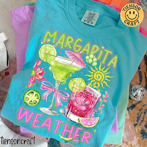 May include: A turquoise Comfort Colors t-shirt with a graphic of a margarita and a pink cocktail. The words "Margarita Weather" are printed on the shirt. The shirt has a smiley face logo that says "Tienson Craft".