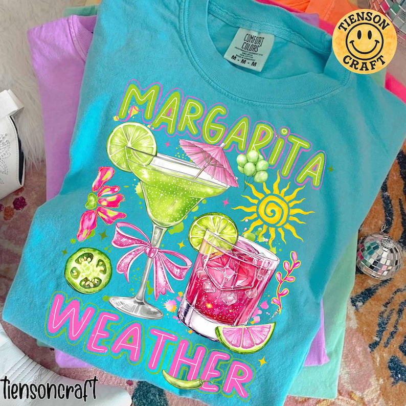May include: A turquoise Comfort Colors t-shirt with a graphic of a margarita and a pink cocktail. The words "Margarita Weather" are printed on the shirt. The shirt has a smiley face logo that says "Tienson Craft".